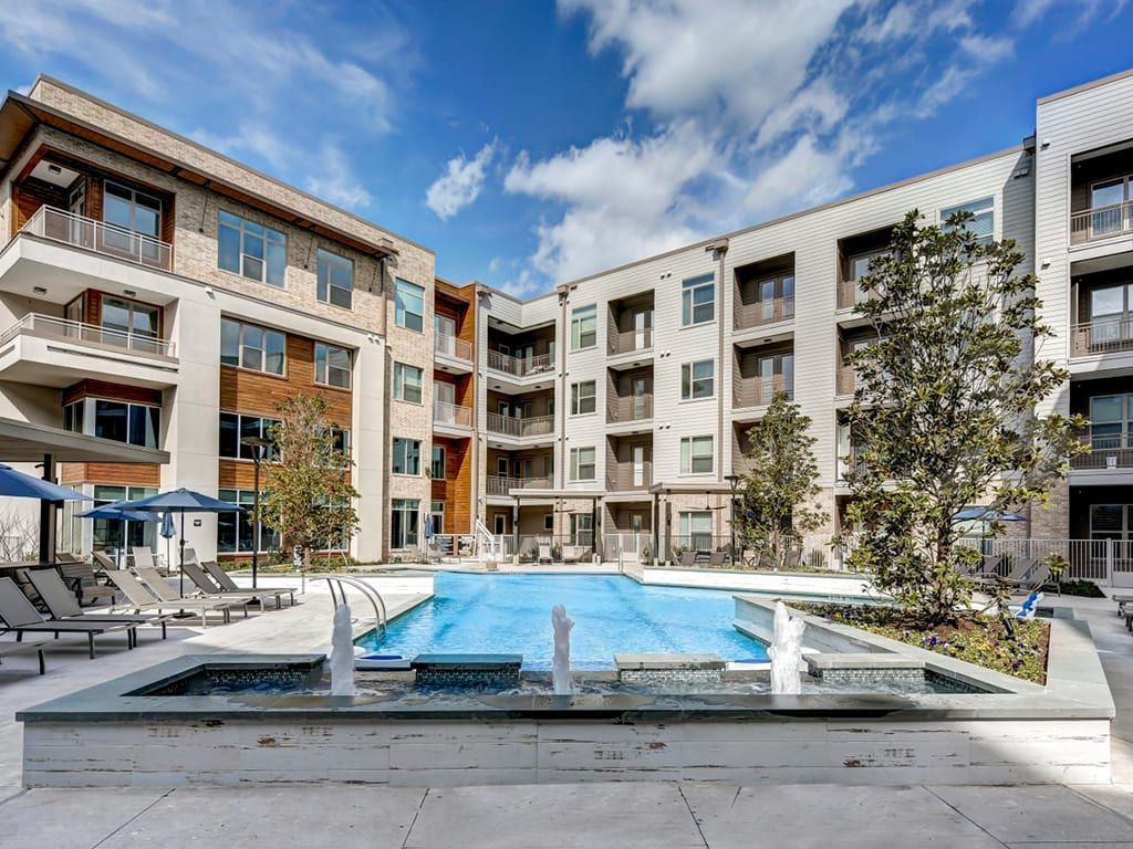 Apartment complex courtyard with a pool, fountains, and lounge chairs under a blue sky.