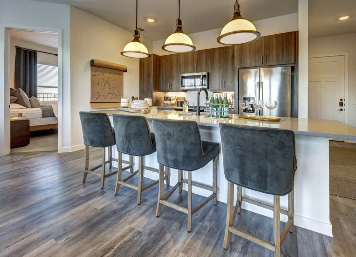 A modern kitchen with a breakfast bar, four gray upholstered stools, and pendant lights. Dark wood cabinets, stainless steel appliances, and wood-look flooring.