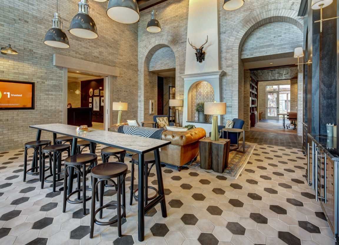 Interior of a lounge area with brick walls, arched doorways, and hexagonal tile floors. A long table with stools is on the left, and couches and a fireplace are in the center.