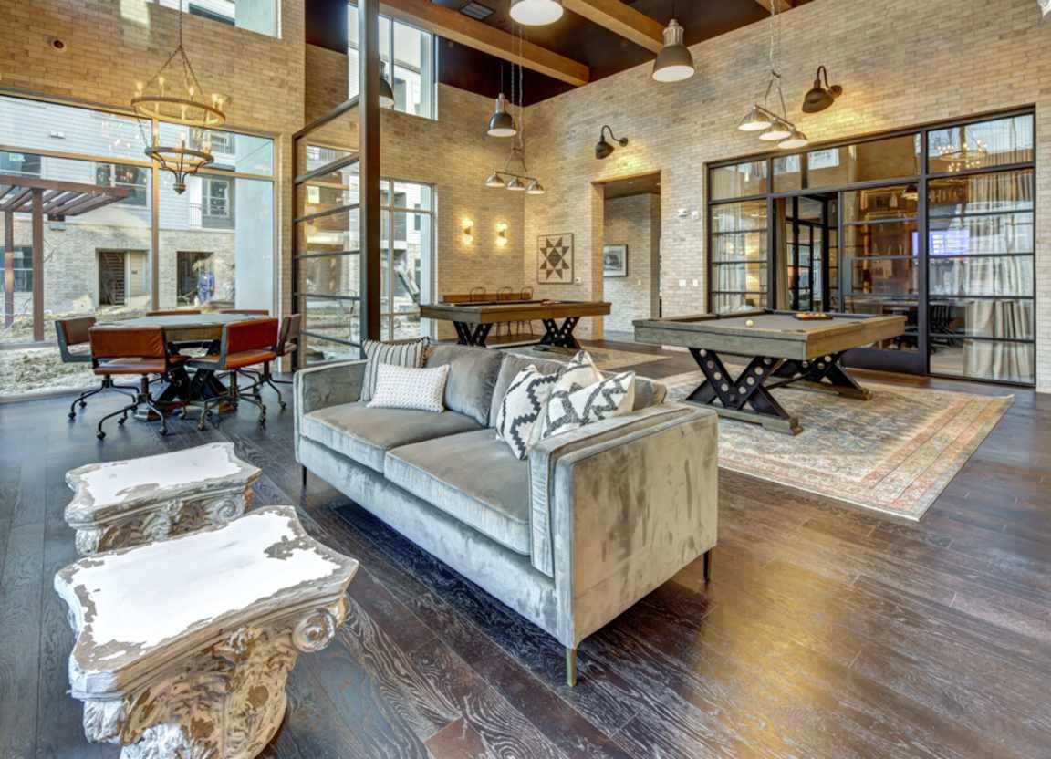 Spacious lounge with a gray sofa, pool tables, and ornate side tables; features exposed brick and dark wood floors.