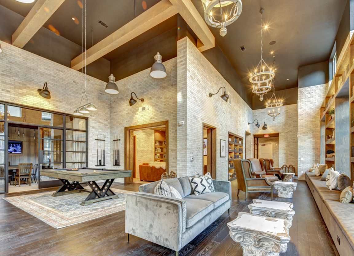 A luxurious lounge area with brick walls, wooden beams, and plush seating. Features include a pool table, library seating, and decorative lighting.