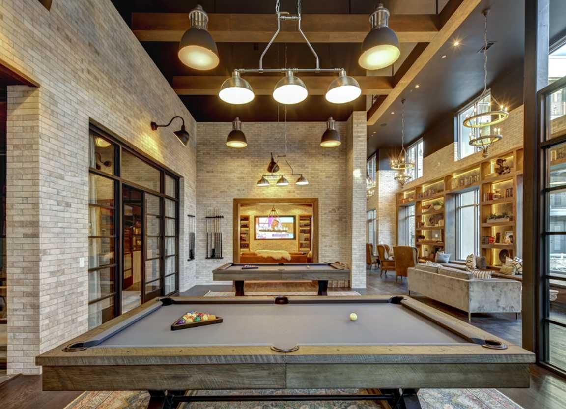 An upscale game room with a pool table, brick walls, and industrial lighting. Large windows offer views of a seating area.