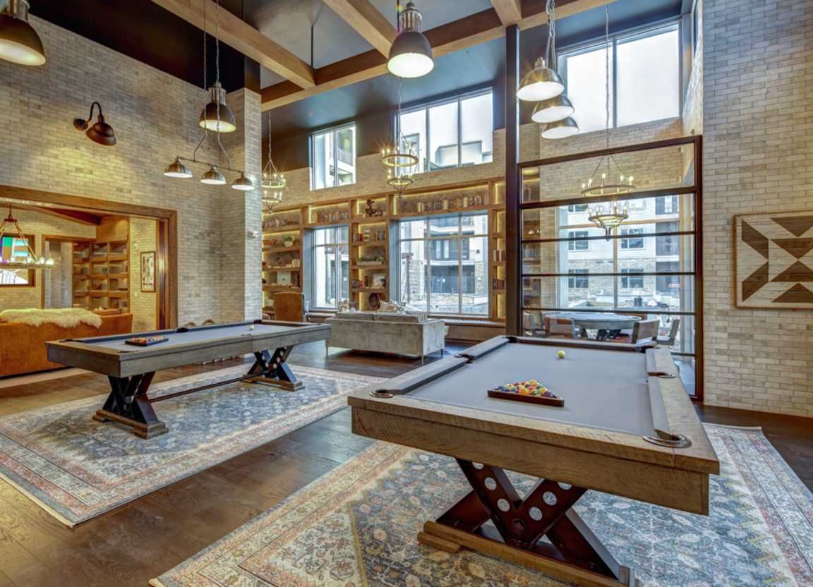 Two pool tables in a spacious room with large windows. Exposed brick walls, wood beams, and pendant lights create a modern, industrial aesthetic.