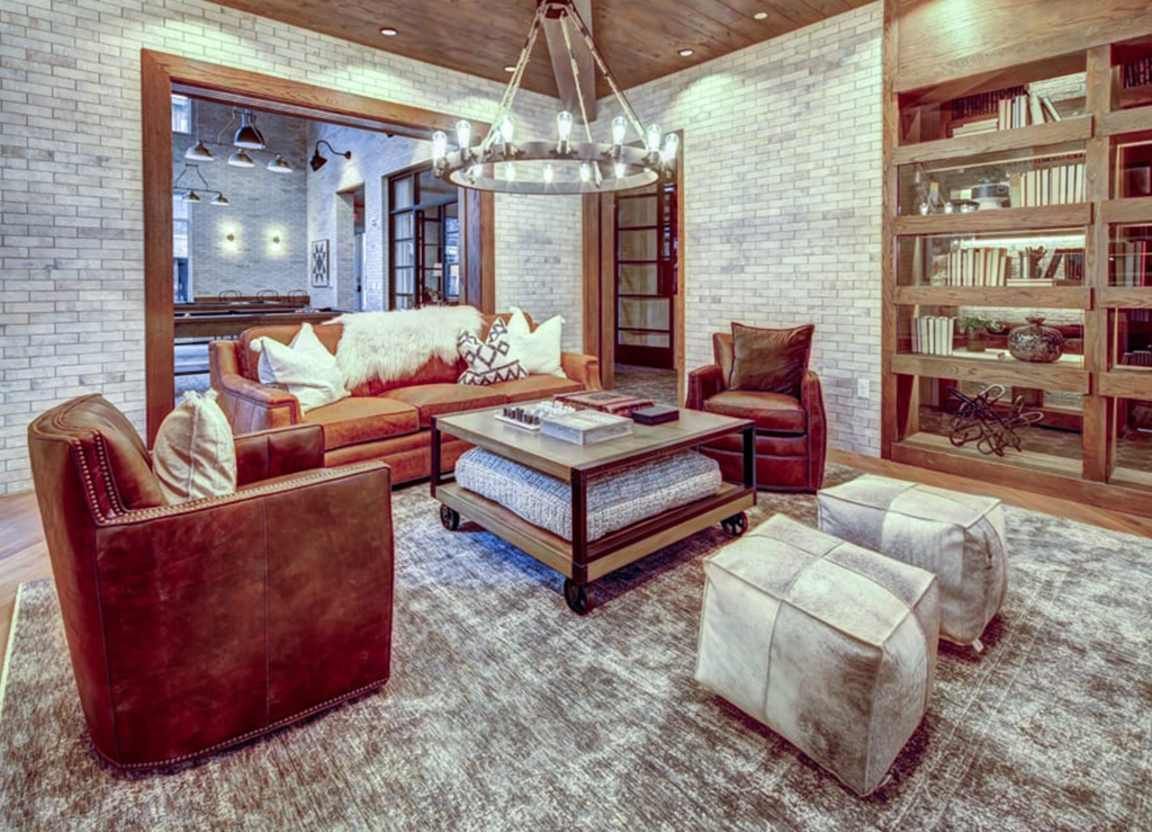 A cozy lounge area with brown leather furniture, a rug, and a large bookcase. White brick walls and a chandelier complete the look.
