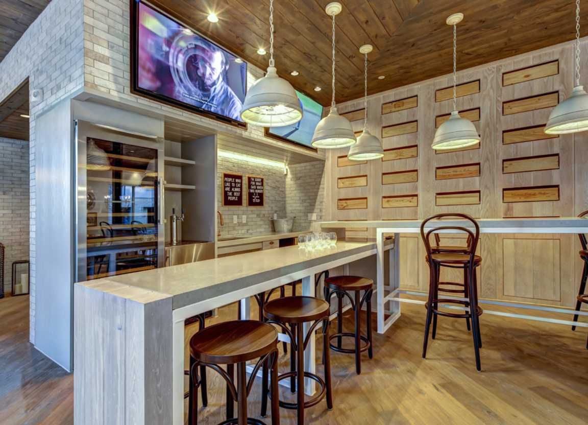 A modern bar area with a wooden bar, stools, wine fridge, and a TV, with pendant lights.
