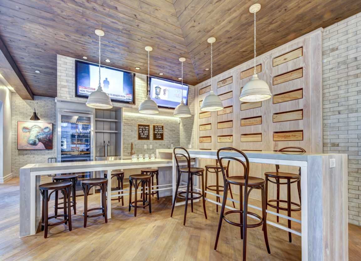 Bar area with wooden accents, pendant lights, TVs, and stools. A light-toned brick wall contrasts with a long bar and various seated stools.