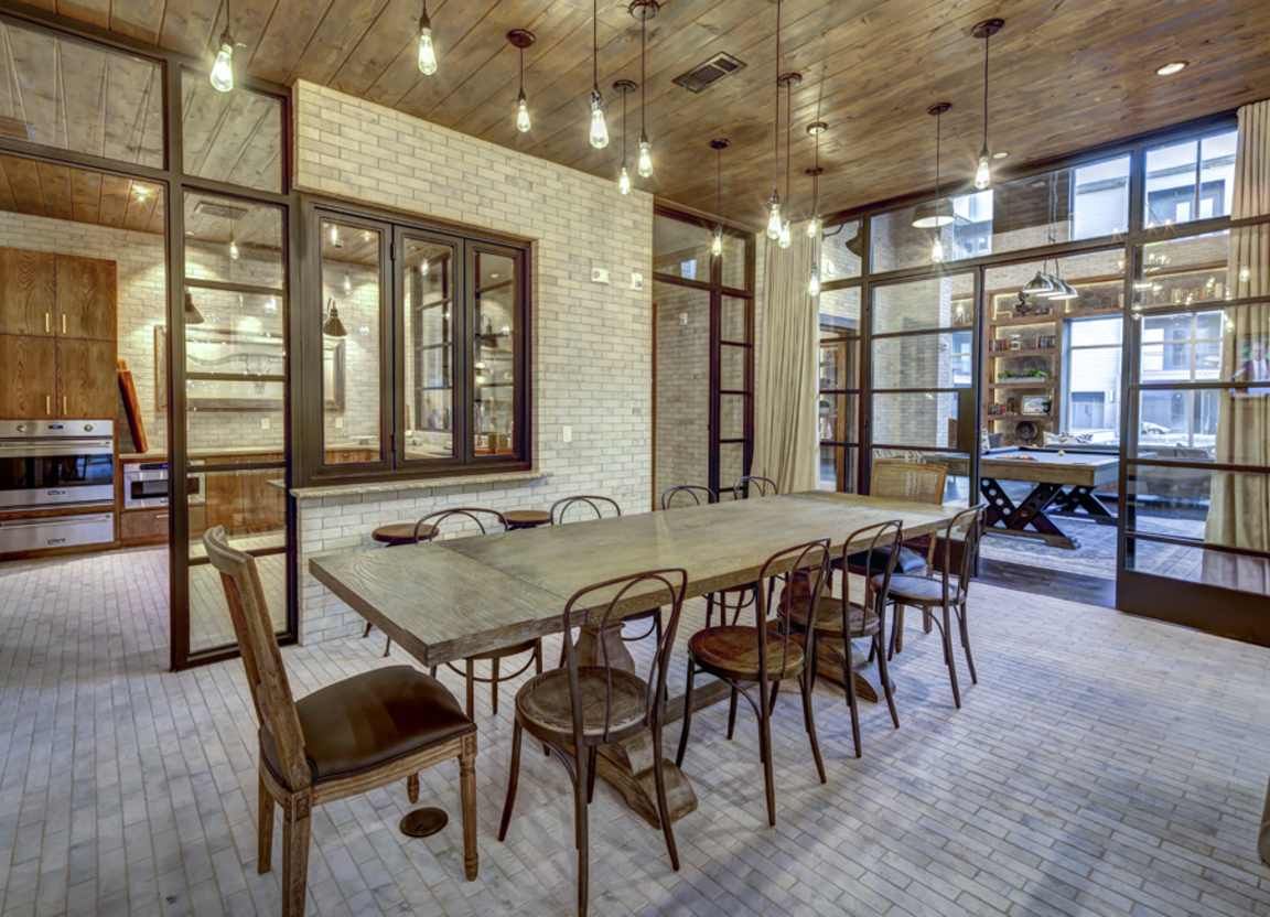 A long wooden dining table with ten chairs, set in a room with brick walls and large windows. Edison-bulb lights hang from the ceiling.
