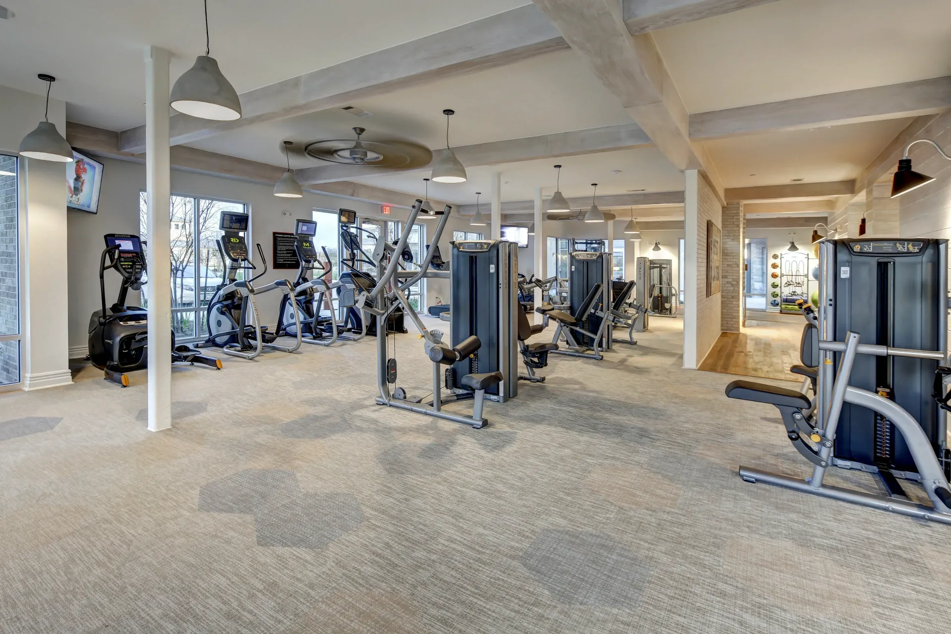A well-lit gym with various exercise machines on speckled carpet. Columns support the exposed beams of the ceiling.