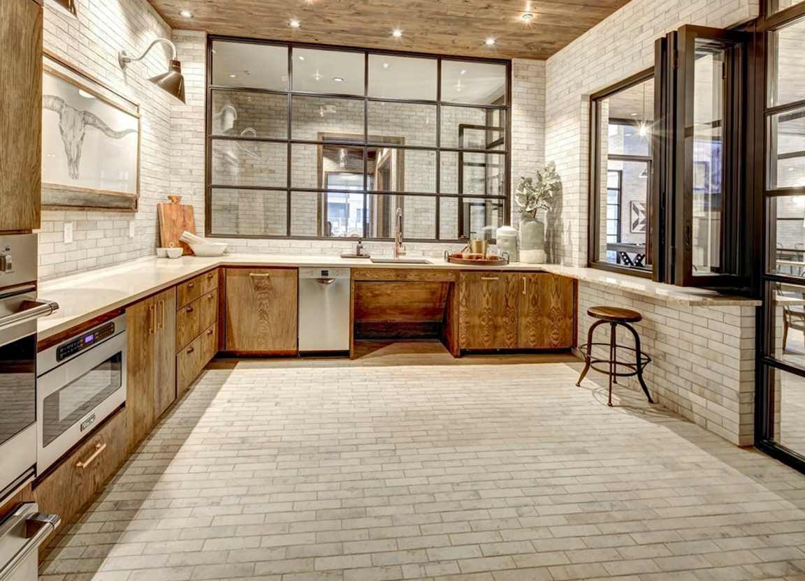 Industrial-style kitchen with white brick walls, wooden cabinets, and a large black-framed window. A stool sits near a small window.