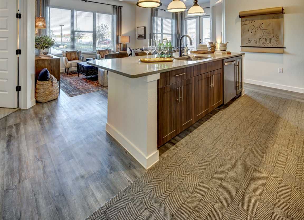 Modern kitchen with a large island, hardwood cabinets, and stainless steel appliances. The living area is visible in the background with large windows.