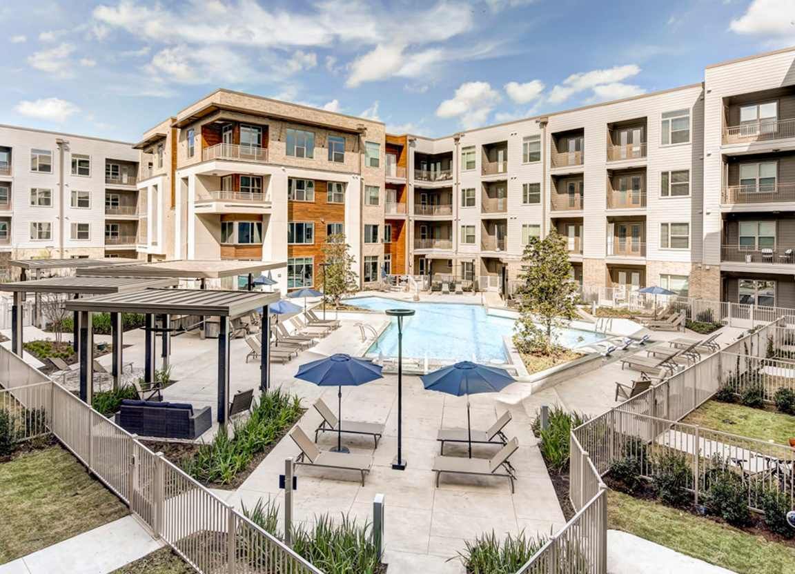 An aerial view of an apartment complex courtyard with a pool, lounge chairs, and umbrellas. The building exterior is beige with multiple stories.