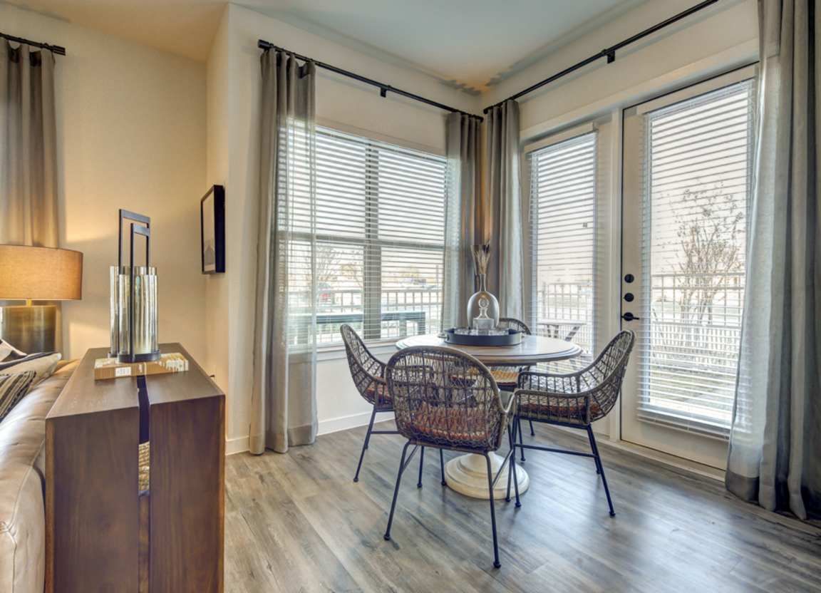 A bright dining area with a round table, four woven chairs, and windows with blinds and curtains.