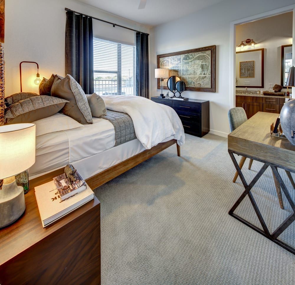 Bedroom with a bed, nightstand, desk, and access to a bathroom. Neutral colors with wood and metal accents create a warm, modern feel.