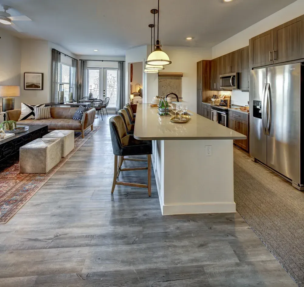 An open-concept living space with a kitchen island, bar stools, and stainless steel refrigerator. The living room has a sofa and rug.