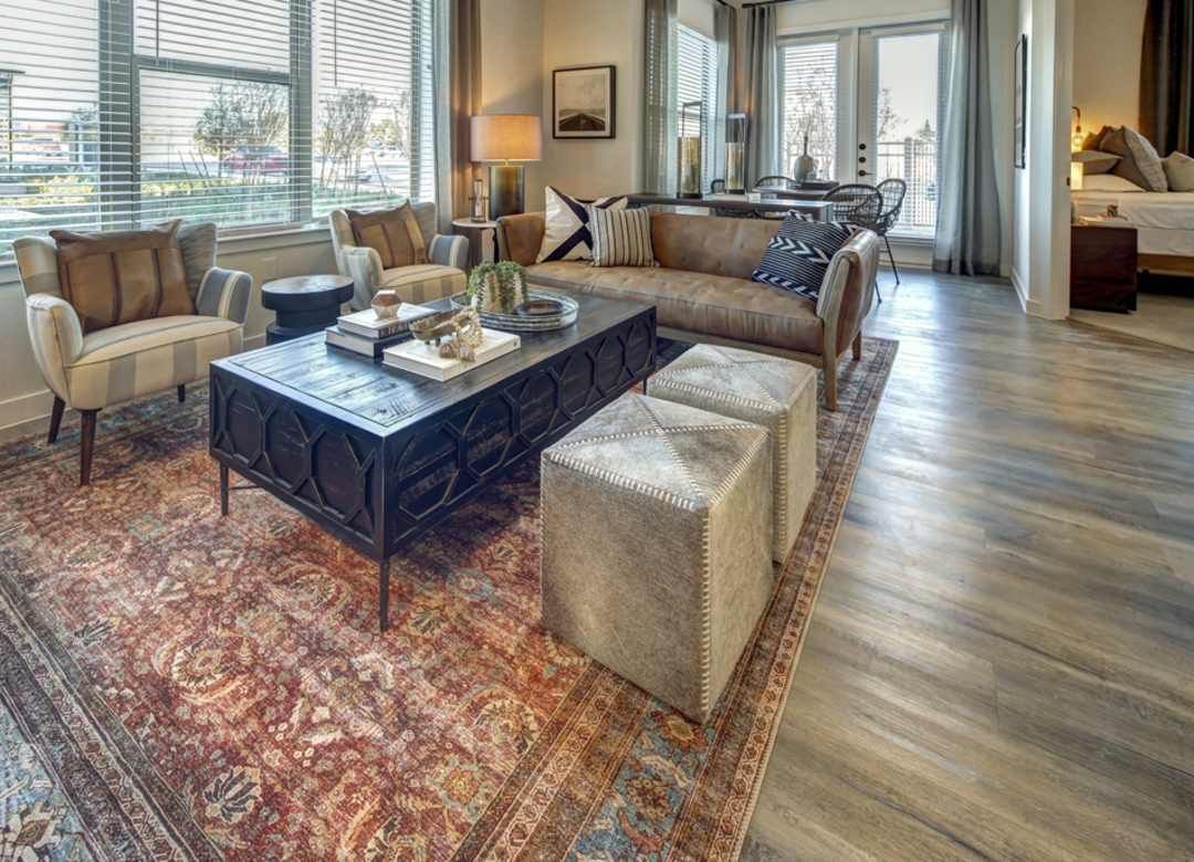 A modern living room with a rug, coffee table, and seating. The room features a window, sofa, and ottoman in a neutral color palette.