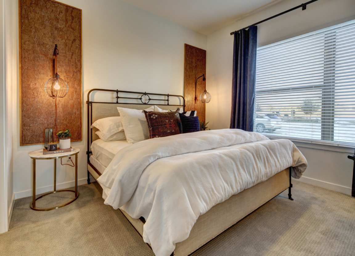 Bedroom with a large bed, nightstand, and window. The room has a neutral color scheme with dark blue curtains and decorative wall panels.