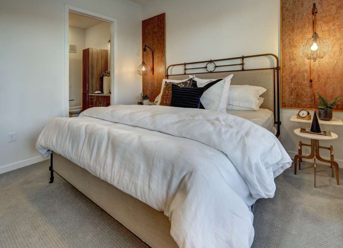 A bedroom featuring a bed with white bedding, a metal headboard, and bedside tables with lights. A wooden wall panel is on the right.