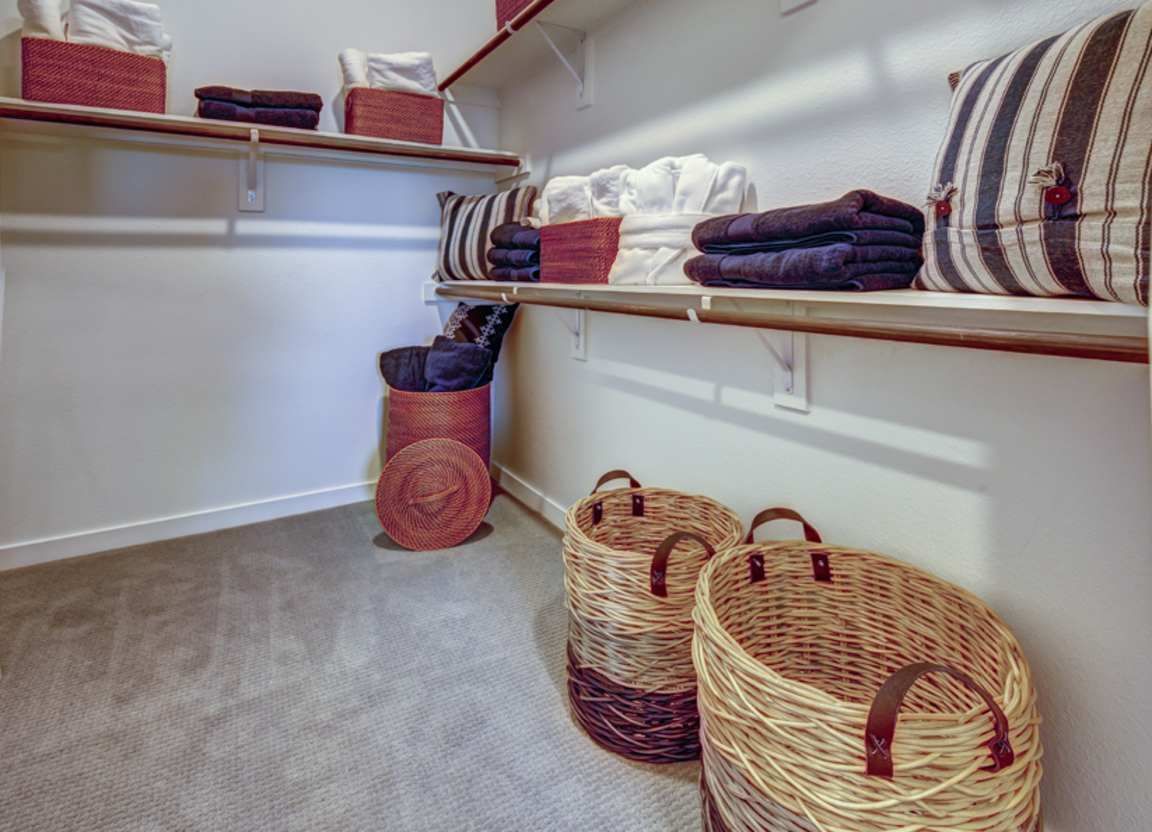 Walk-in closet with shelves holding folded towels, baskets, and pillows. Light wood shelves against white walls and gray carpet.