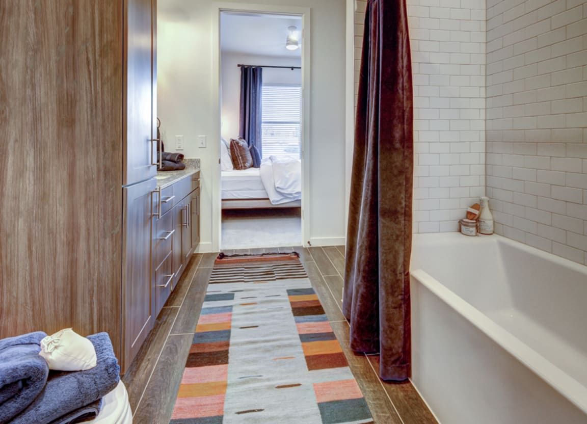 A bathroom hallway with a rug, wooden cabinets, a bathtub, and a partially visible bedroom.