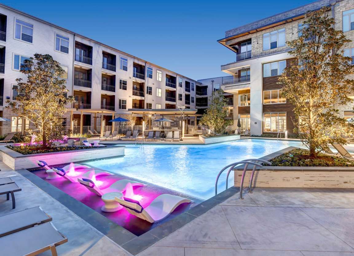 A luxurious apartment complex courtyard at dusk. Features a pool with illuminated lounge chairs and buildings with lights.