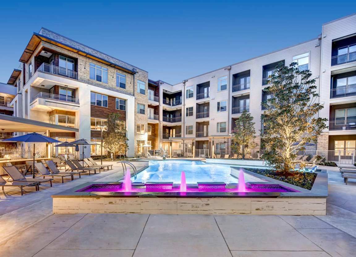 Apartment complex courtyard with a pool illuminated by pink lights, surrounded by lounge chairs and buildings.