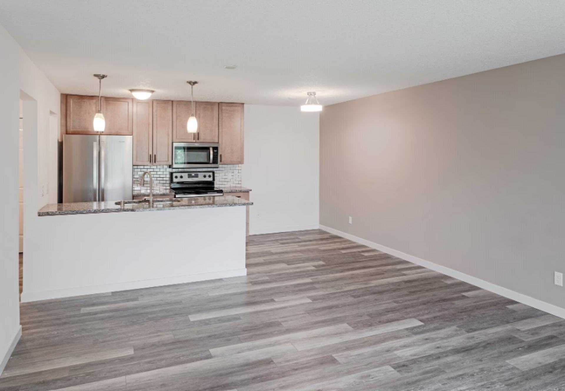 An empty living room with hardwood floors and a kitchen in the background.