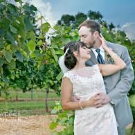 Couple in formal attire embracing outdoors by a leafy tree, sharing an intimate kiss.