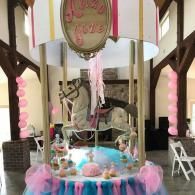 Decorated indoor birthday display with pink streamers, balloons, a cake table, and a carousel horse centerpiece