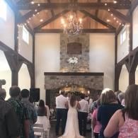 Guests seated in a rustic wedding hall as the bride and groom stand before a stone fireplace altar
