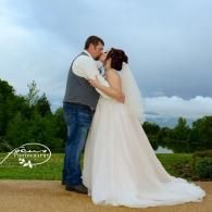 Bride and groom embracing outdoors on a cloudy day, with a flowing white wedding dress and blue sky behind them
