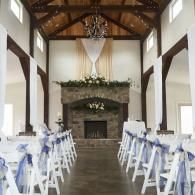 Wedding ceremony aisle in a rustic chapel with white chairs, blue ribbons, and a stone fireplace altar