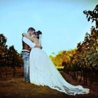 Couple embracing in a vineyard at sunset, with a long white dress trailing behind.