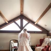 Person in a white dress standing in a sunlit attic room near a window and upholstered chair
