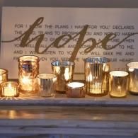 Glass votive candles glowing on a shelf in front of a framed wall quote.