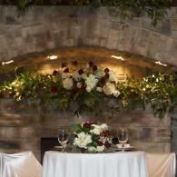 Floral wedding arch on stone wall above a decorated table with white cloth and candles