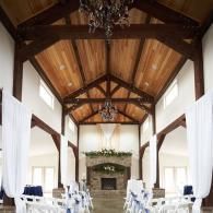 Elegant banquet hall with high wooden ceiling, chandeliers, white drapes, and stone fireplace.
