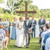 Wedding couple walking down aisle outdoors, guests seated on either side, with a large wooden cross behind them