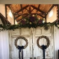 Rustic wedding backdrop with white doors, greenery, wreaths, and floral arrangement under exposed wooden beams