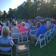Outdoor gathering with people seated on white folding chairs on a grassy lawn at sunset