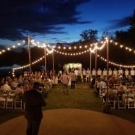 Outdoor wedding reception under string lights at dusk, with guests seated along a central aisle.