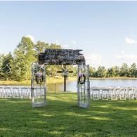 Outdoor wedding arch by a lake with white chairs on a grassy lawn under a bright blue sky