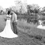 Couple in wedding attire standing by a calm pond in a grassy field