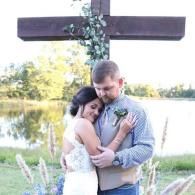Couple embracing outdoors beside a wooden cross by a lake during sunset