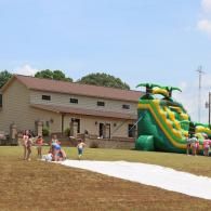 Outdoor scene with a green inflatable slide beside a beige building, with children gathered on a grassy field.