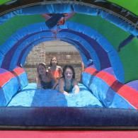 Three people sliding inside a blue-and-red inflatable tunnel at an outdoor park