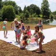 Children playing on a giant white tarp outdoors near a pond, using colorful water balloons