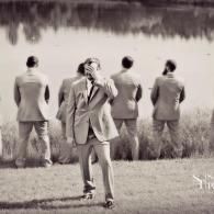 Four men in suits stand and look over a lake, one facing the camera in the foreground.