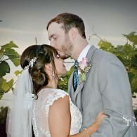 Bride and groom embracing and kissing at an outdoor wedding, with flowers and greenery in the background