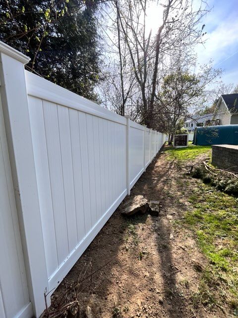 White vinyl fence along a narrow dirt path beside a yard with trees and a rock.