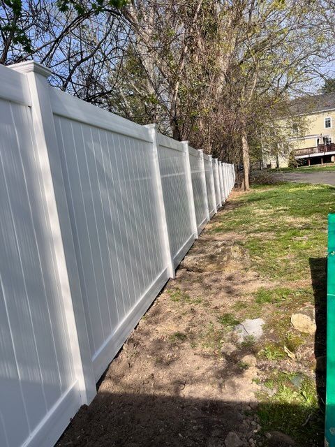 White vinyl fence along a narrow grassy yard with trees and a house in the background